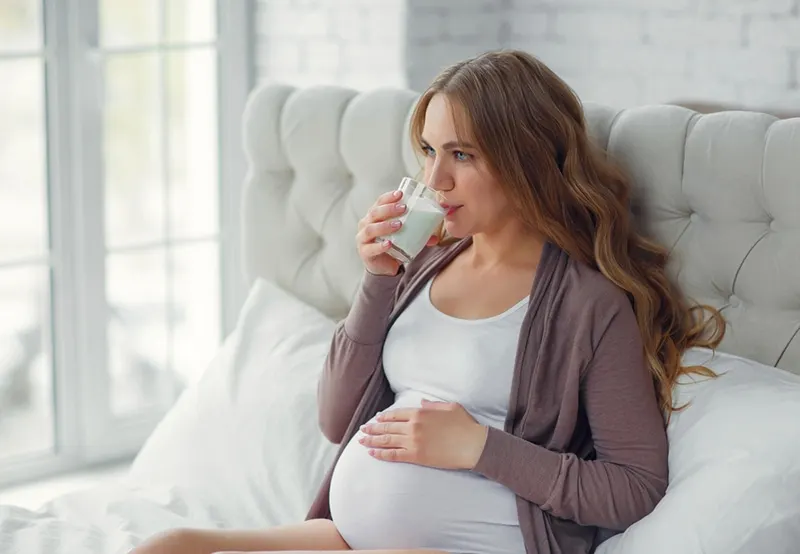 Schwangere trinkt Milch, während sie auf dem Bett sitzt
