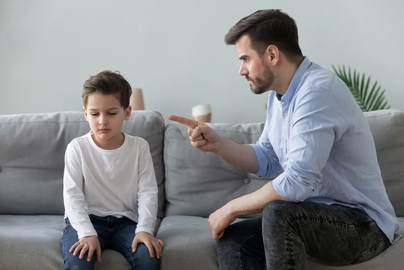 Wütender Vater schimpft mit dem traurigen Jungen, der neben ihm auf der Couch sitzt
