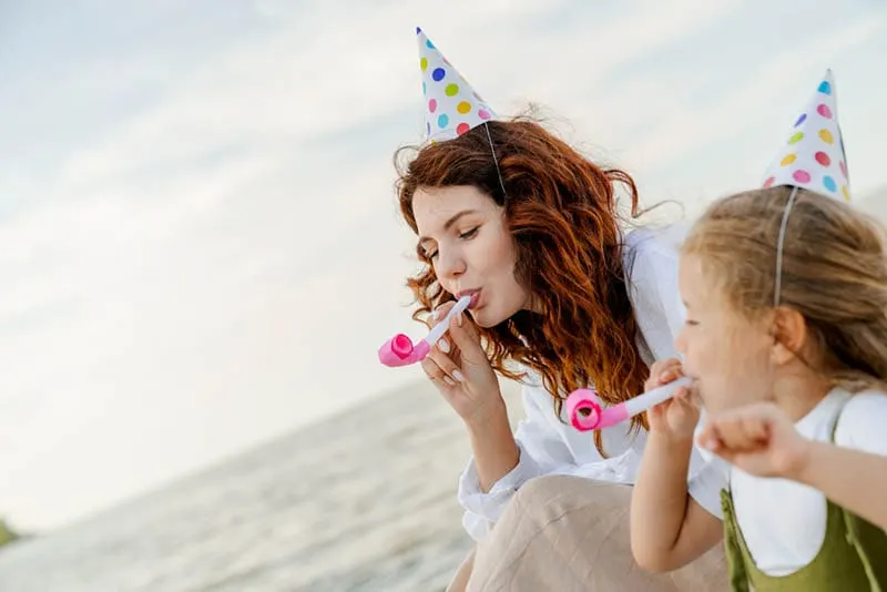 eine Tante und ein kleines Mädchen mit Partyhüten sitzen am Strand und blasen in Pfeifen