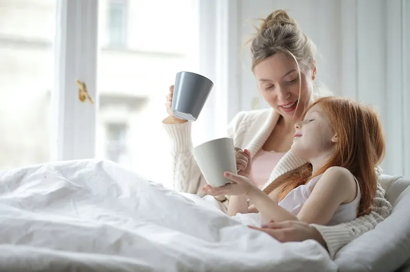 fröhliche Frau, die mit Tochter spricht, während sie Tee in einem Bett trinkt