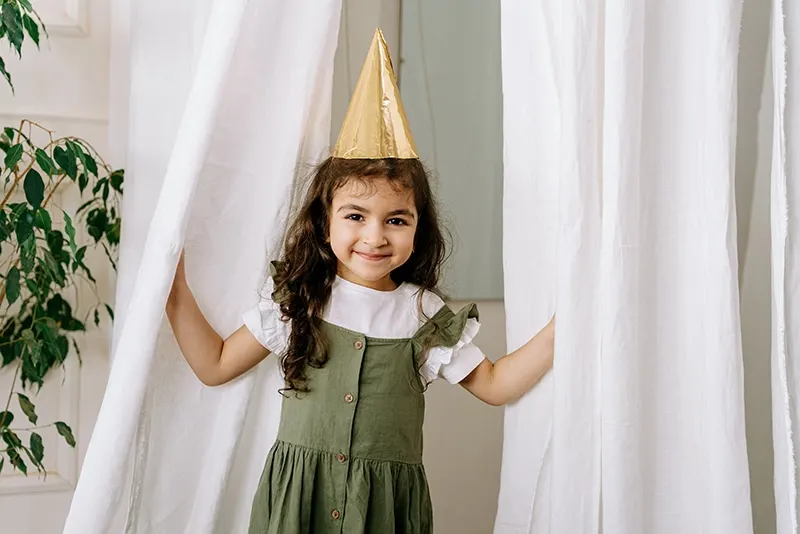 kleines Mädchen mit Partyhut, das zwischen Vorhängen steht
