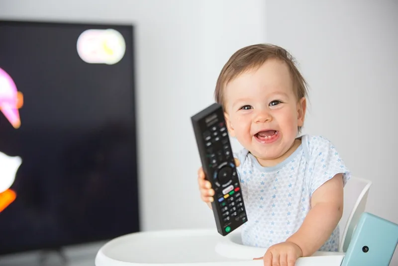 lächelndes Baby, das eine Fernbedienung hält, während es vor dem Fernseher sitzt
