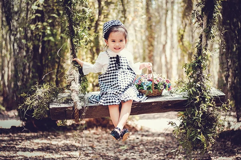 lächelndes kleines Mädchen, das auf der Schaukel sitzt und einen Korb mit Blumen hält