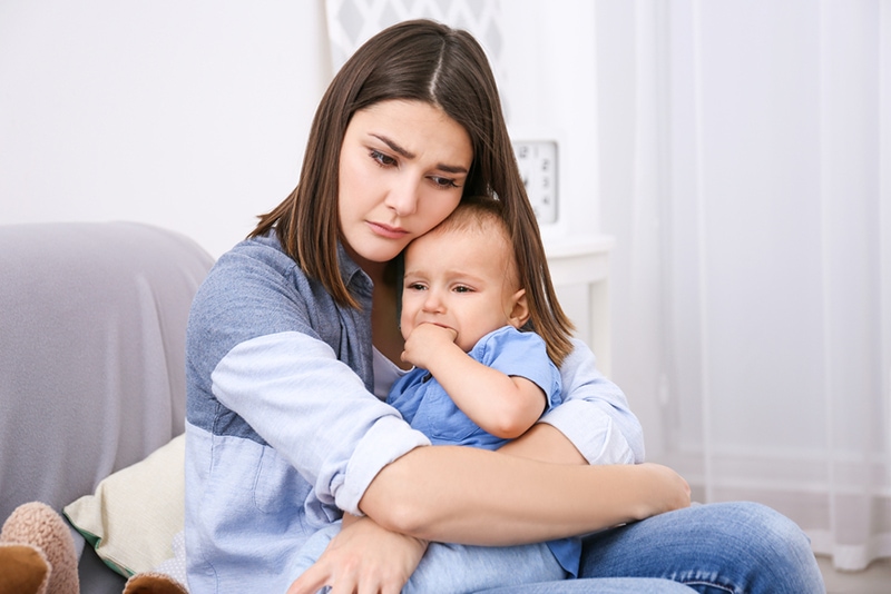 Depressive junge Frau umarmt ihr Baby, während sie auf der Couch sitzt