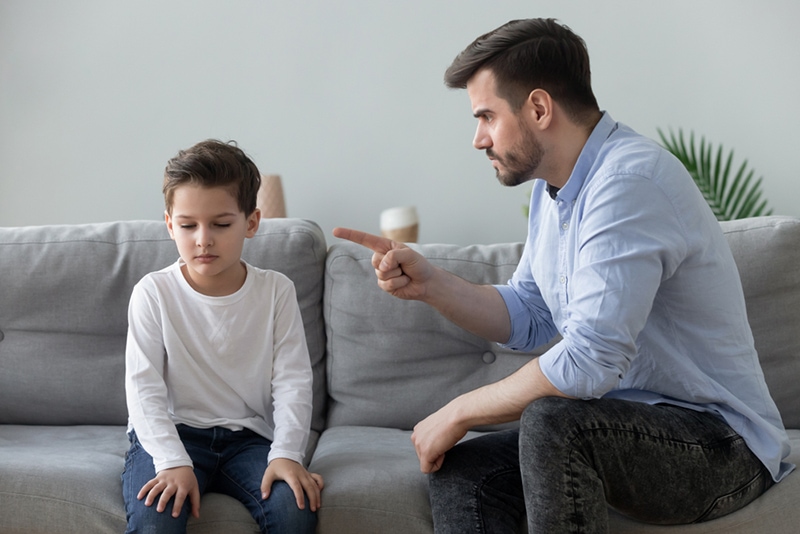Wütender Vater schimpft mit einem traurigen Jungen, der auf der Couch sitzt