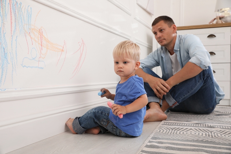 kleiner Junge, der zu Hause an die Wand zeichnet, während sein Vater ihn ansieht