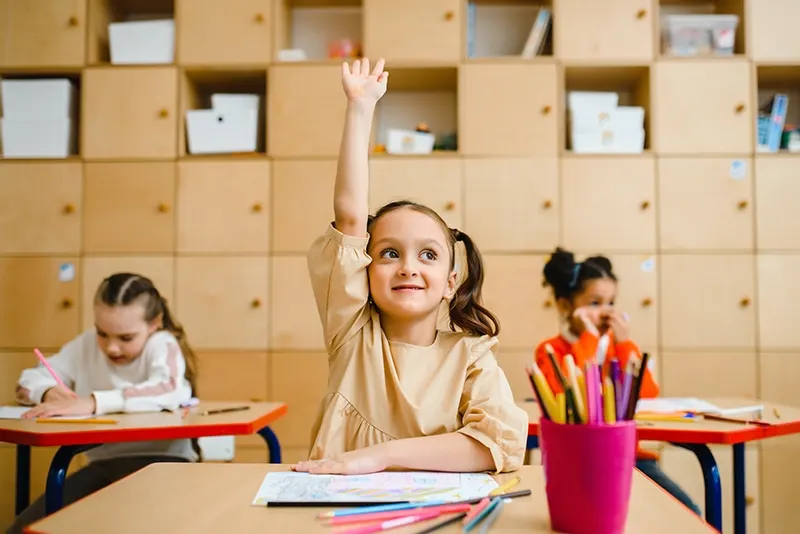 kleines Mädchen, das im Klassenzimmer die Hand hebt, während es auf dem Schreibtisch sitzt