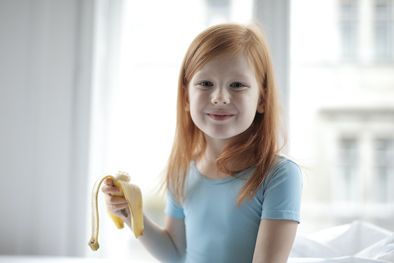 kleines Mädchen mit roten Haaren, das eine Banane hält und lächelt