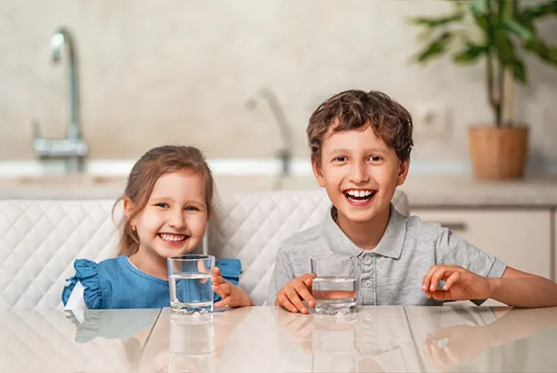 lächelndes Mädchen und Junge, die ein Glas mit Wasser halten, das im Esszimmer sitzt