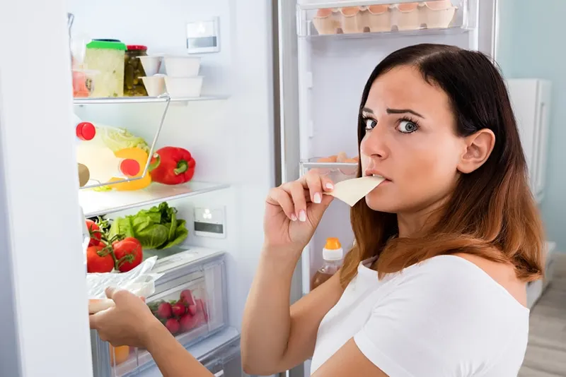 verwirrte Frau isst Käsescheibe vor offenem Kühlschrank