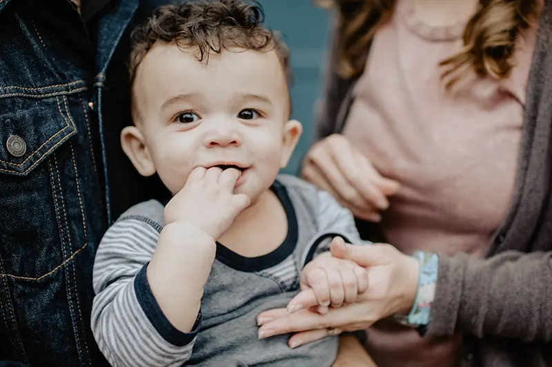 Baby, das in die Finger beißt, während seine Eltern ihn tragen