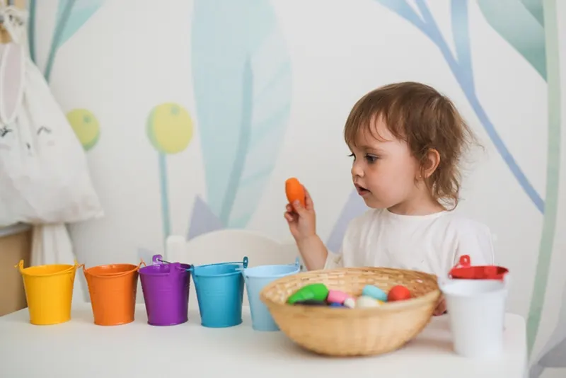 Kleinkindmädchen sortiert buntes Spielzeug nach farbigen Eimern am Tisch