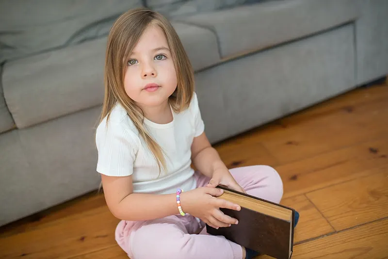 kleines Mädchen, das ein Buch hält und auf dem Boden sitzt