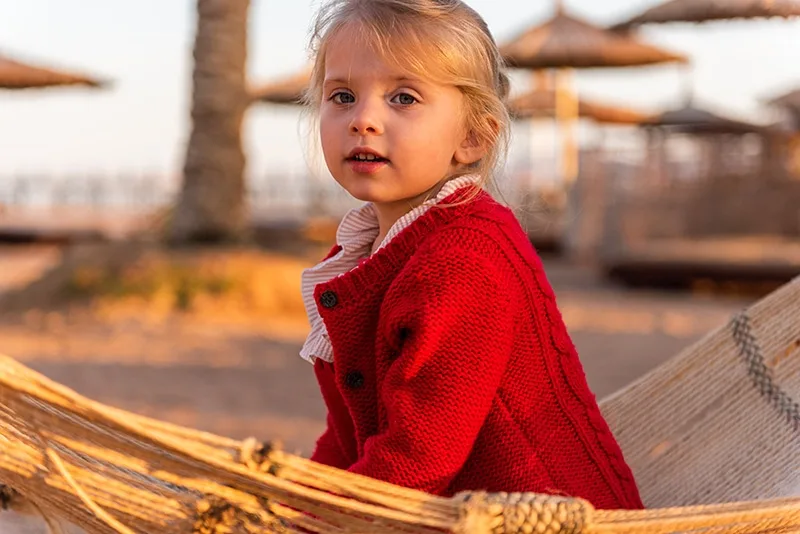 kleines Mädchen im roten Pullover sitzt auf einer Hängematte am Strand