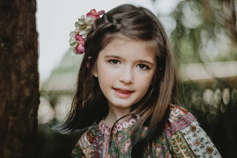 kleines Mädchen mit Blumen im Haar, das in der Nähe des Baumes steht