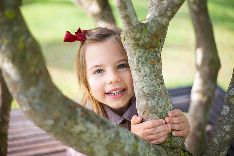 kleines Mädchen mit roter Schleife im Haar versteckt sich hinter dem Baum
