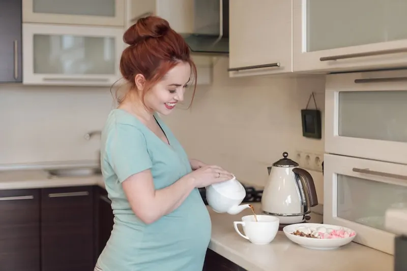 Schwangere gießt Tee aus einer weißen Teekanne in eine weiße Tasse