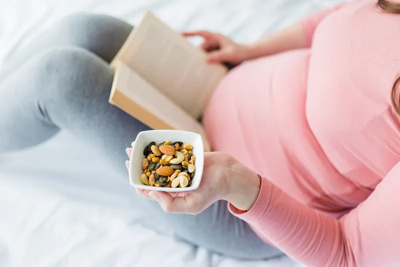 Schwangere Frau, die auf dem Bett sitzt und Nüsse isst, während sie ein Buch liest