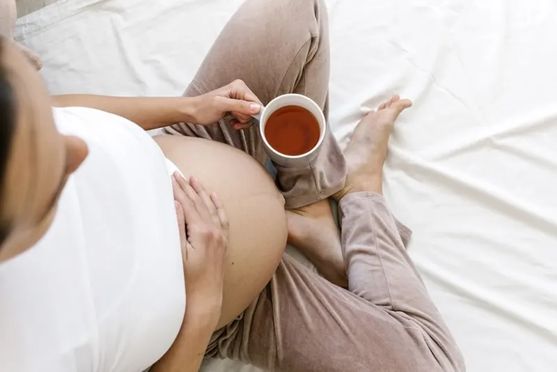 Schwangere trinkt Tee, während sie auf dem Bett sitzt