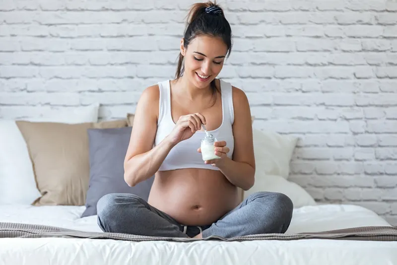 junge schwangere frau isst joghurt, während sie zu hause auf dem bett sitzt