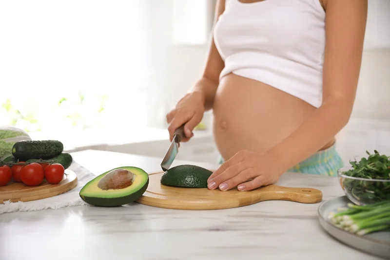 Schwangere Frau schneidet Avocado am Tisch in der Küche