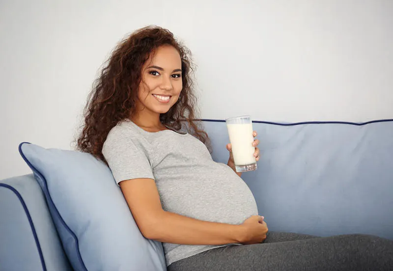 glückliche schwangere Frau auf der Couch sitzend mit einem Glas Milch