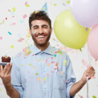 lächelnder mann, der geburtstag mit luftballons und muffin feiert