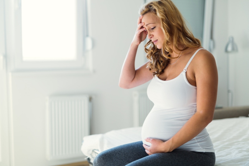 Junge schwangere Frau mit Schmerzen auf dem Bett sitzend