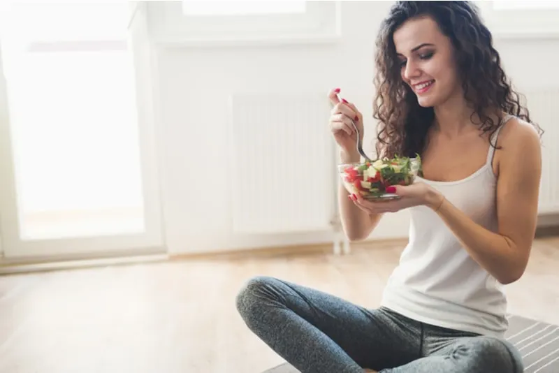 lächelnde Frau, die auf dem Boden sitzt und einen Salat isst