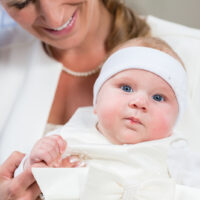 Patin hält Baby, das ein weißes Taufkleid trägt