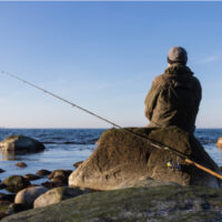 Rückansicht eines Meerforellenanglers mit Spinnrute auf einem Felsen sitzend