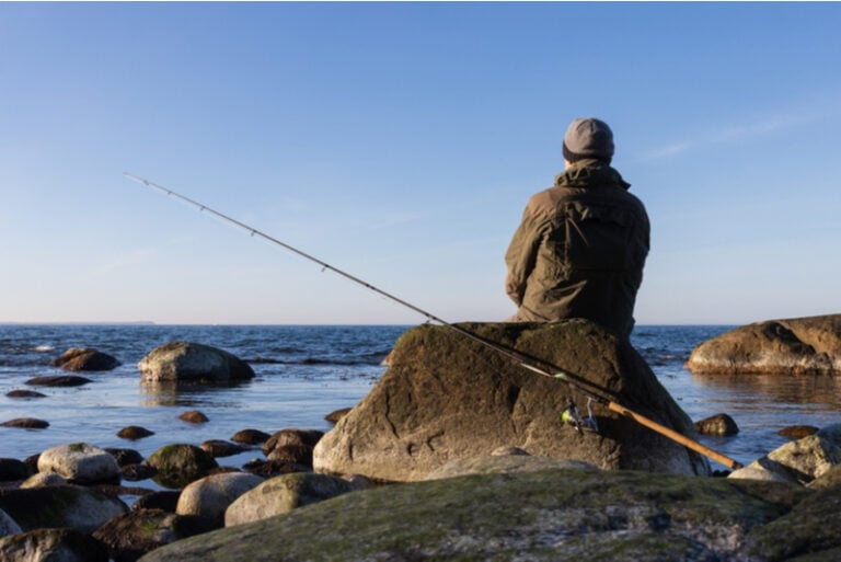 Rückansicht eines Meerforellenanglers mit Spinnrute auf einem Felsen sitzend
